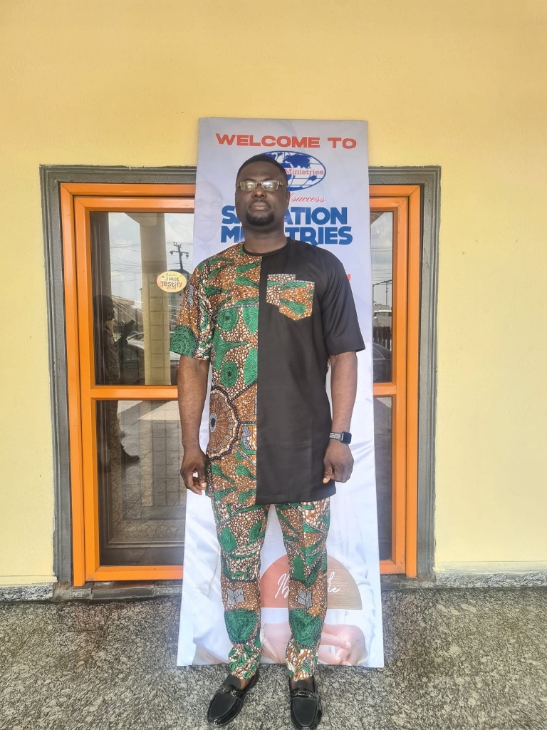 Uche Okochi standing in front of a Salvation Ministries Warri Delta banner.